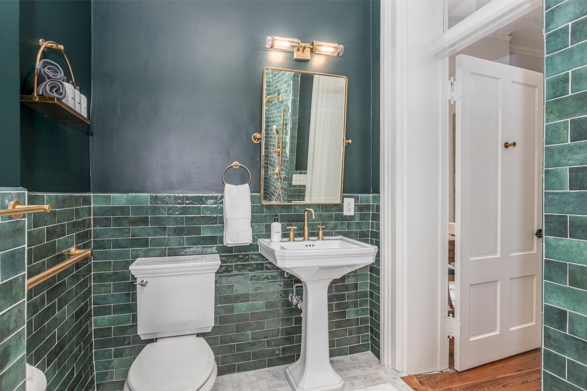 A bathroom with green tiled walls, a white toilet, a pedestal sink, a mirror, a towel rack with a white towel, and shelves holding rolled towels and soap.