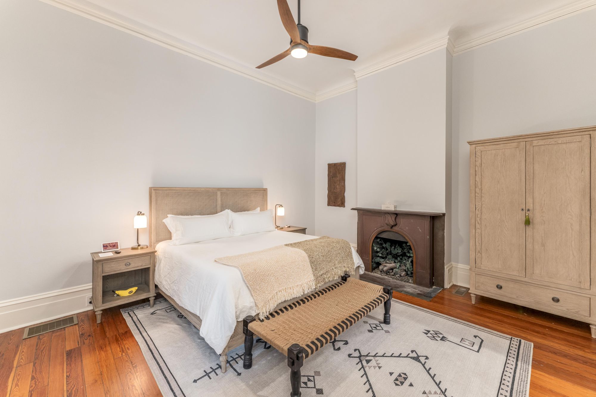 A well-lit bedroom with a bed, two side tables, an armchair, a fireplace, a wardrobe, a bench, and a ceiling fan on a white ceiling.