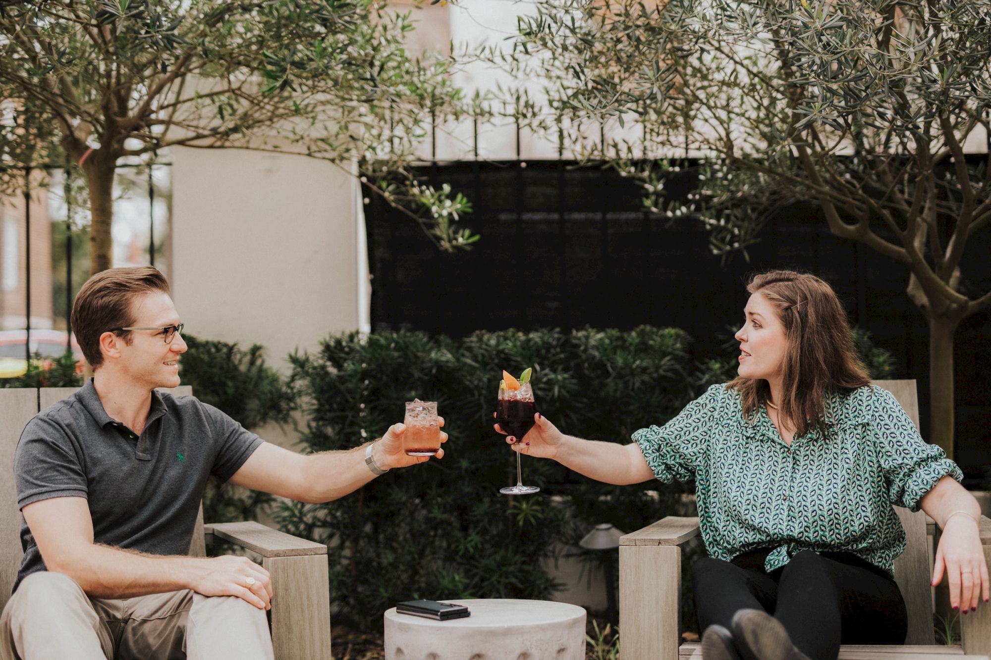 Two people sitting outdoors, raising their drinks for a toast, surrounded by greenery and trees.