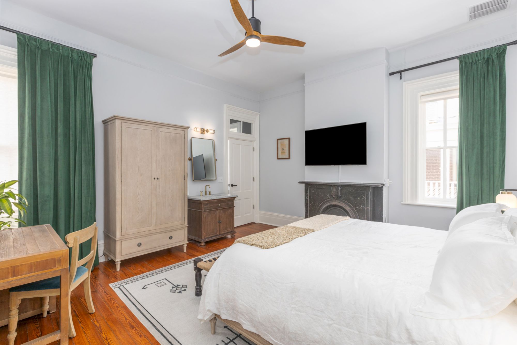 A bright bedroom features a white bed, wooden furniture, green curtains, a TV, and ceiling fan. The room is well-lit by natural light coming through windows.