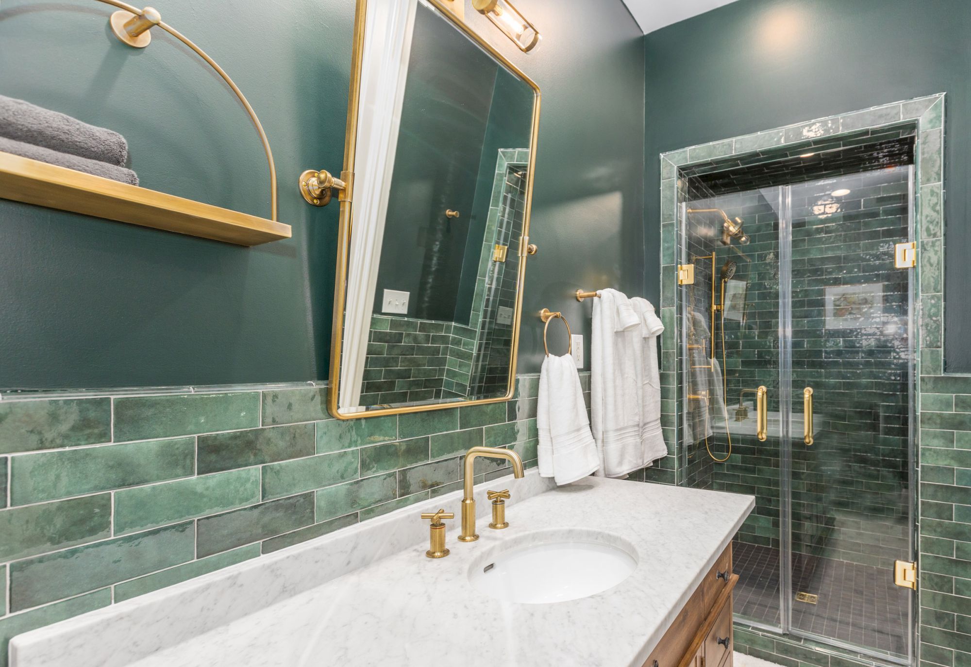 A modern bathroom with green subway tiles, gold fixtures, a marble countertop, a large mirror, and a glass-enclosed shower with towels hanging nearby.