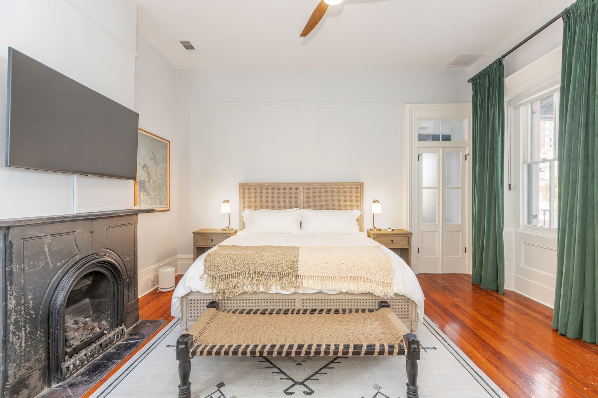 A cozy bedroom with a bed, wooden floors, a fireplace, a TV, green curtains, and nightstands on each side. White walls and large windows end the sentence.