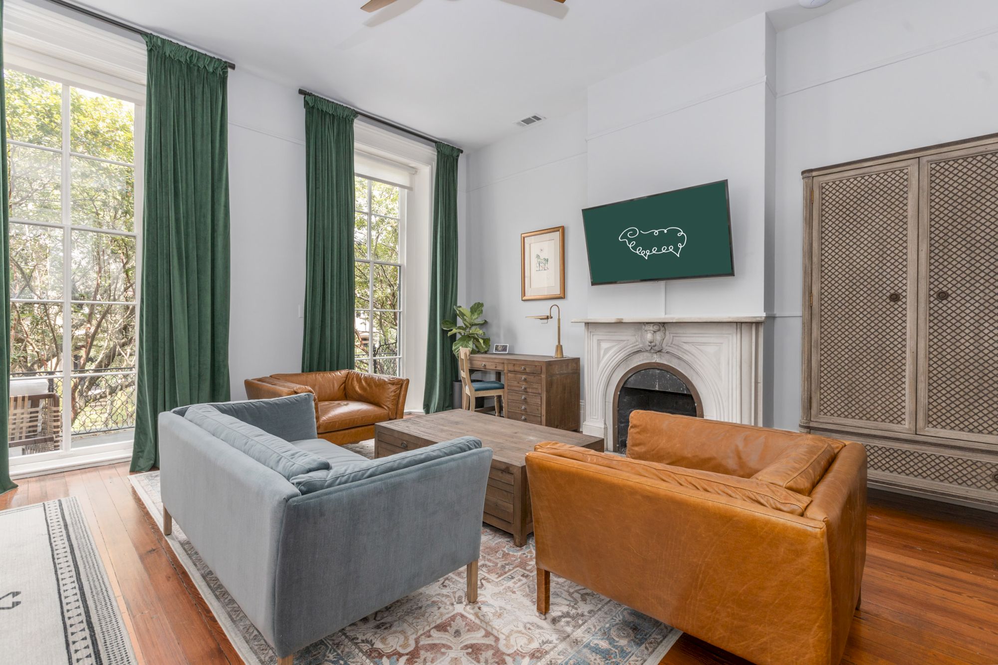 A stylish living room with two brown leather chairs, a gray couch, green curtains, a large window, a TV above a fireplace, and wooden furniture.