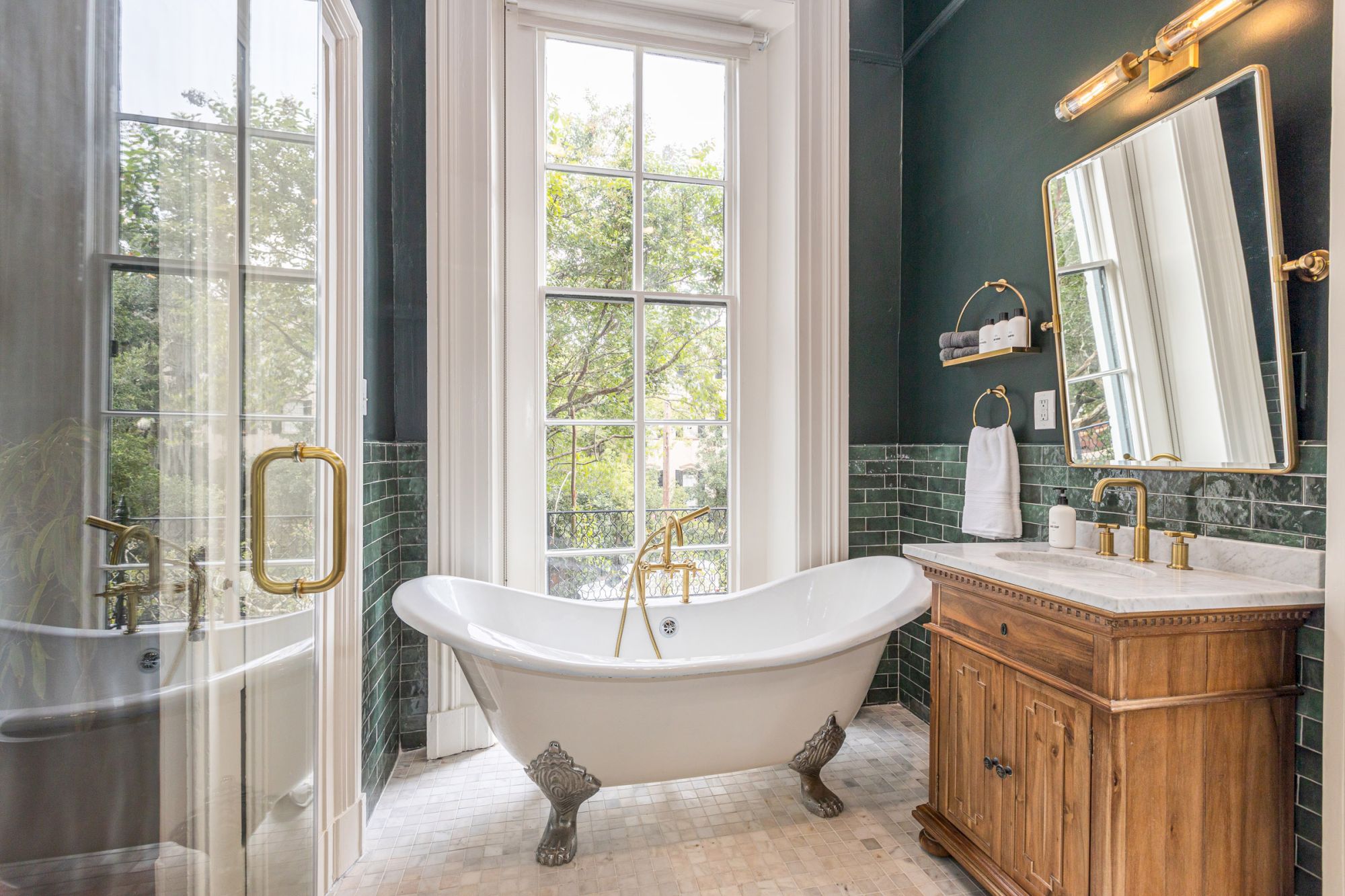 A bathroom with a clawfoot bathtub, wooden vanity, large window, green tiles, and gold fixtures. There is a towel on the wall and a mirror above.