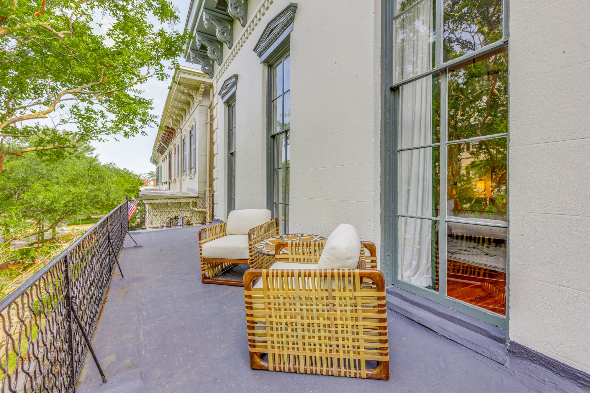 The image shows a balcony with two wicker chairs and cushions near a large window, surrounded by trees and adjacent to a building with ornate design.