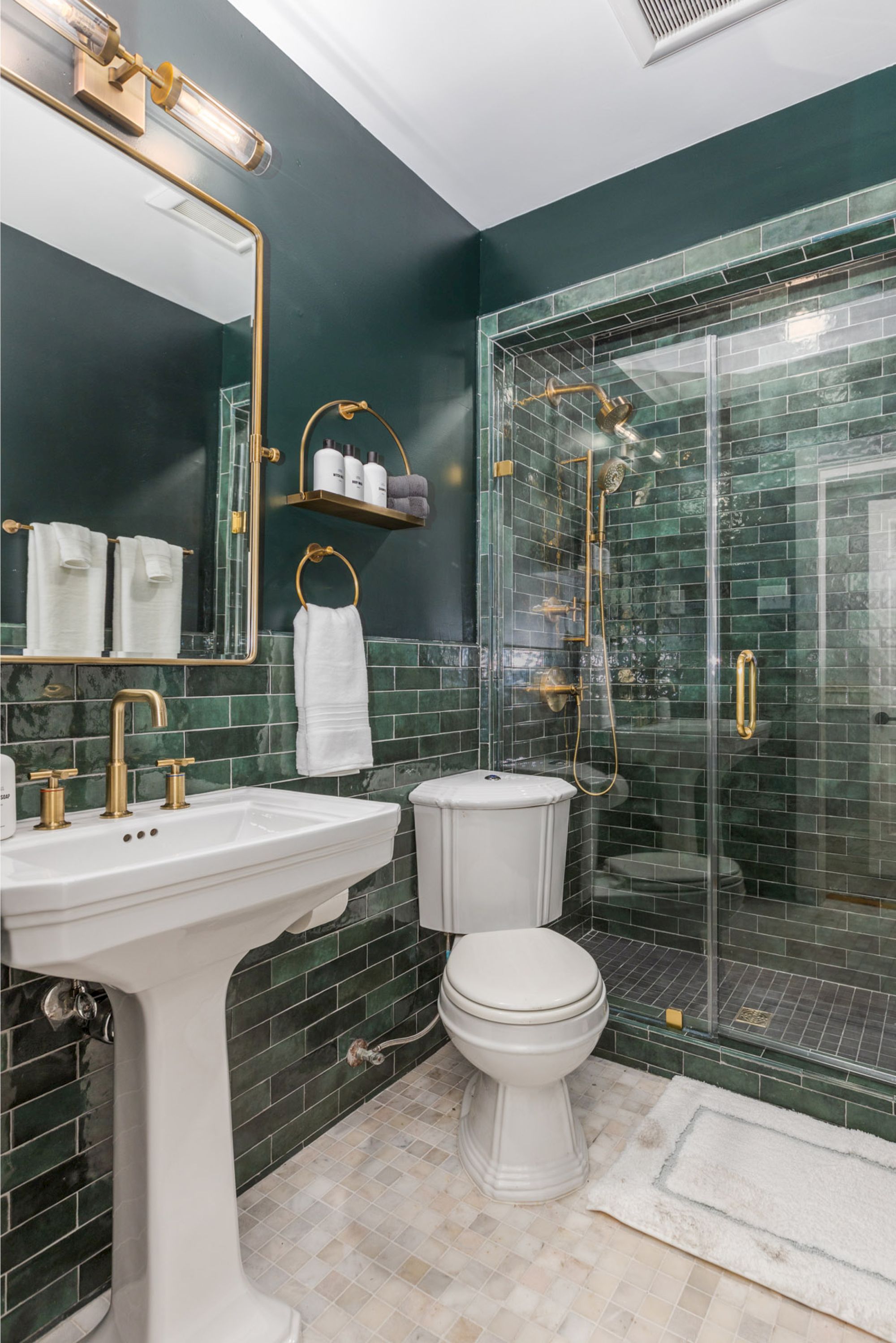 A modern bathroom with dark green tiles, a glass shower, a pedestal sink, a toilet, and a mirror above the sink.