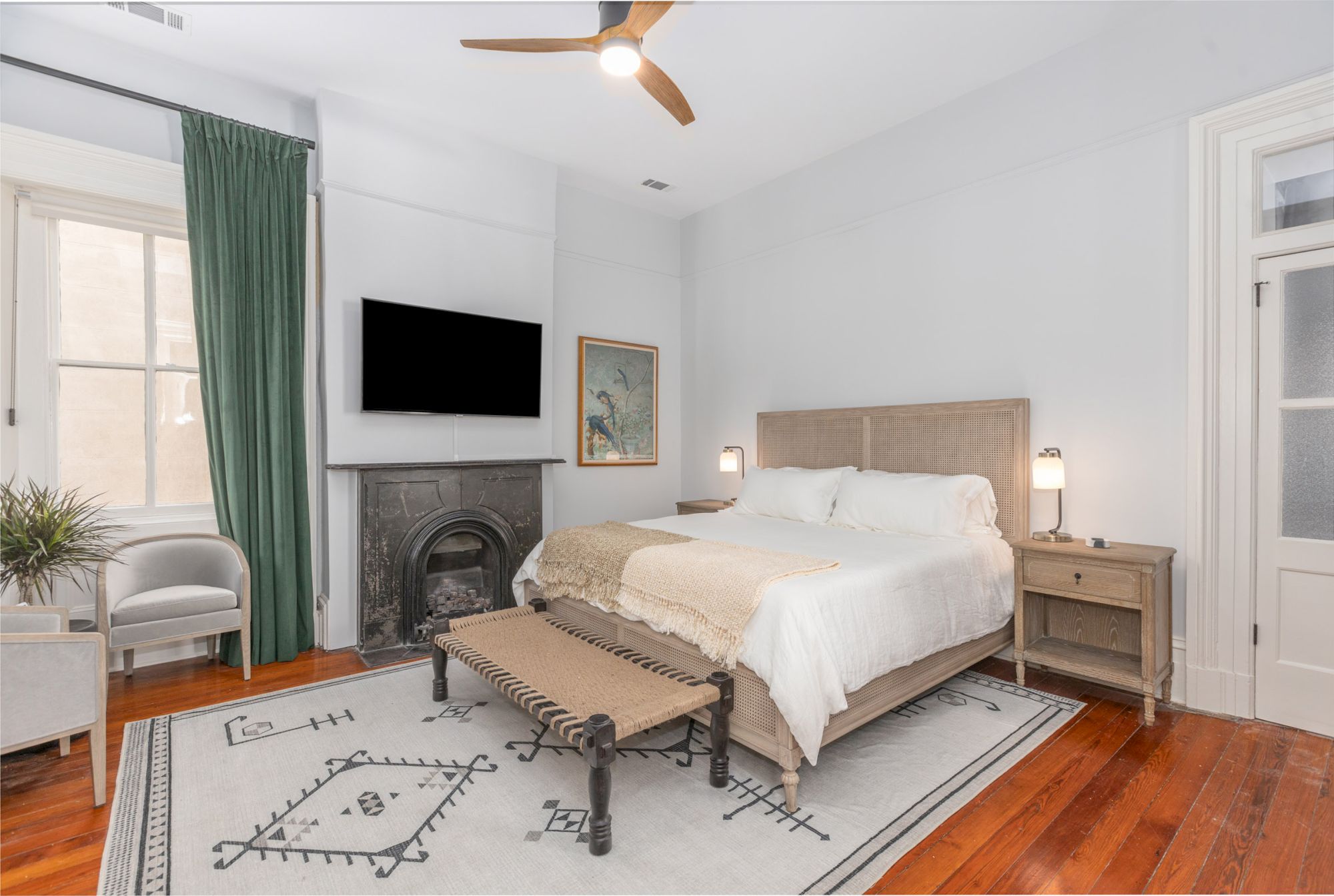 A bedroom with a bed, side tables, a TV, a fireplace, a bench, a potted plant, a chair, and a green curtain. The room has wooden flooring and a rug.