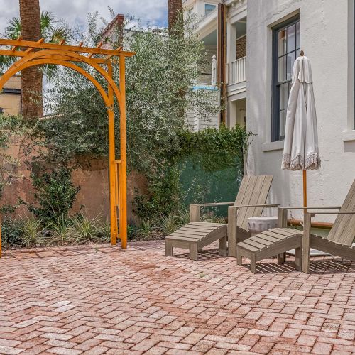 The image shows a backyard patio with two wooden lounge chairs, a closed umbrella, and a wooden trellis against a white house.