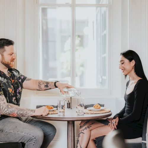 A man with tattoos and a woman sit at a round table in a bright room, with the man pouring water into a glass while they both smile.