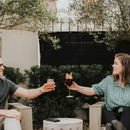 Two people sitting outside, clinking glasses in a toast while smiling at each other, surrounded by greenery and outdoor furniture.