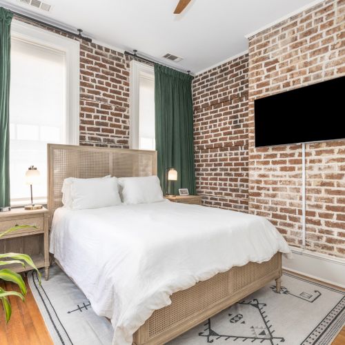 A cozy bedroom with a bed, white bedding, brick accent wall, wall-mounted TV, green curtains, side table, lamp, rug, and a potted plant.