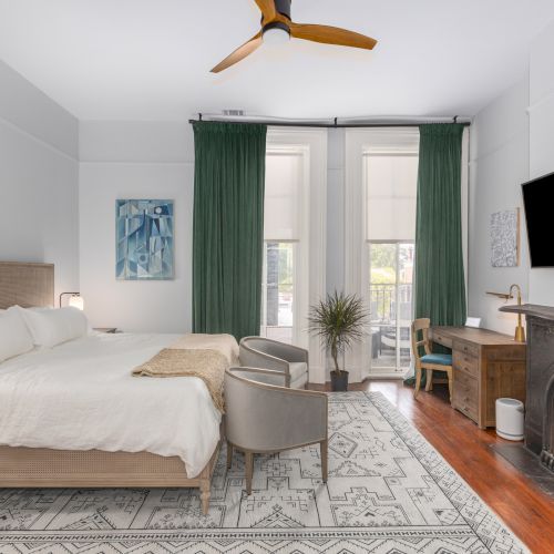 A modern bedroom with a large bed, two gray chairs, green curtains, a wall-mounted TV, a desk, and a fireplace, all in a bright, well-lit space.