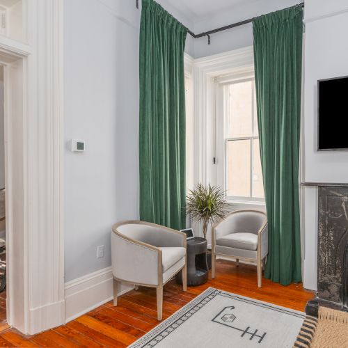 A cozy room with two chairs, a small table, green curtains, a TV mounted above a fireplace, and a rug. An adjacent office space is visible.