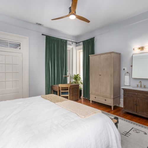 A bedroom with a bed, desk by green curtains, ceiling fan, wardrobe, and vanity with mirror on a wooden floor, lit by wall sconce.