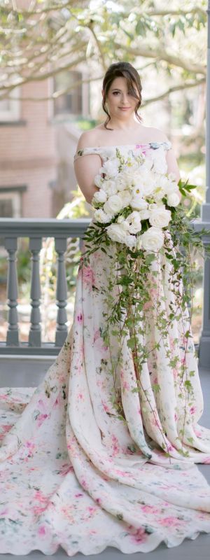 A woman in a floral wedding gown stands on a porch holding a large cascading bouquet of white flowers and greenery, next to a decorative wrought iron railing.