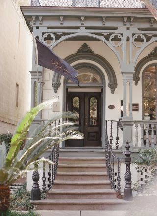 The image shows the entrance of a building, featuring ornate architectural details, a staircase, potted plants, and a flag mounted on the wall.