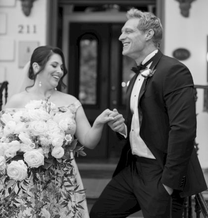 A smiling couple, with the man in a suit and the woman holding a bouquet of flowers, stands in front of a building entrance marked 