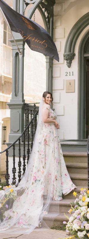 A woman in a floral wedding dress stands on stairs outside a building with black railings, flowers, and a flag that reads 