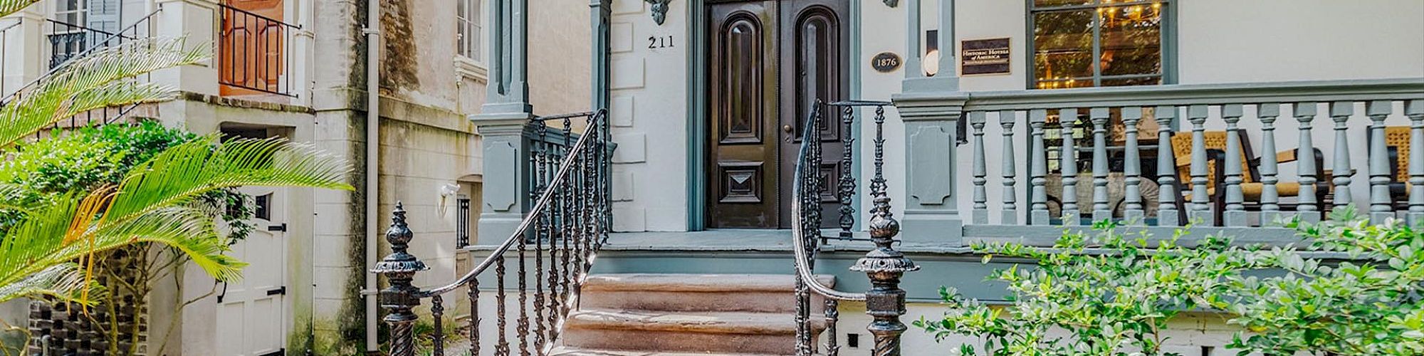 A charming historic house entrance with a detailed arched doorway, ornate railings, and lush greenery surrounding the steps leading to the porch.