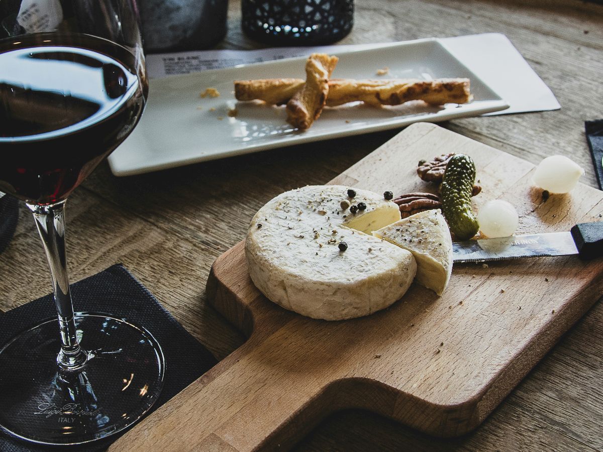 A glass of red wine, a cheese wheel with a slice cut out, pickled vegetables, and some breadsticks on a wooden table.